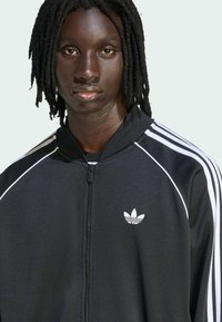 Μαύρο τζάκετ σπιτάκι με φερμουάρ και λευκές ρίγες στα μανίκια, με μικρό λευκό λογότυπο Adidas στην αριστερή πλευρά. Λείο, ελαφρύ ύφασμα.