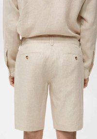Beige linen shorts with a straight cut, featuring two back pockets with button closures and a flat waistband. Smooth texture throughout.