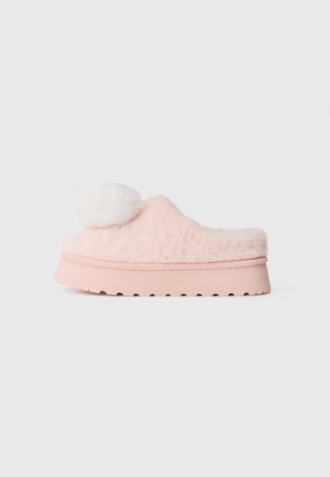 Chausson rose en peluche avec un détail en pompon blanc, une texture douce et une semelle en caoutchouc rose épaisse avec un motif en vague sur le bord.