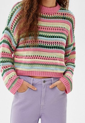 Persoon draagt een meerkleurige gestreepte gebreide trui met open weefsel, gecombineerd met hoge taille lavendelkleurige broek, handen in de zakken.