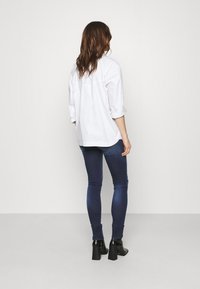 Camisa blanca de botones con mangas remangadas, combinada con jeans ajustados de color azul oscuro y botines negros de tacón, vista desde la espalda.