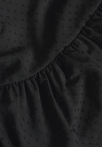 Svart tyg med en texturerad mönster, som har små upphöjda prickar. Materialet har en mjuk, matt yta och rynkade detaljer längs kanten.