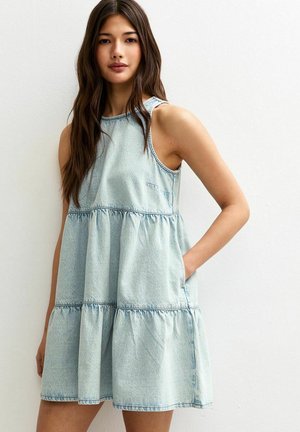 Vestido de mezclilla azul claro con un diseño en capas, cuello redondo y bolsillos. Presenta un corte relajado y detalles de costura visibles.