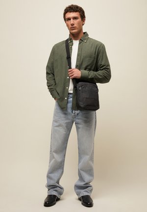 Noor mees, kandes rohelist ruudulist särki, valget t-särki, helesiniseid teksapükse, musti saapaid, hoides musta ristihoidmisega kotti neutraalse taustaga.