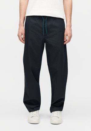 Hombre con pantalones negros holgados con cordón color verde azulado y zapatillas blancas, de pie contra un fondo claro y liso.