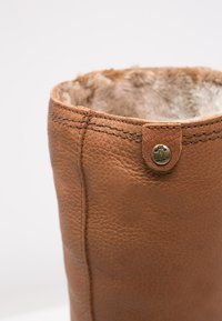 Bota de couro castanho com um forro interior peludo, apresentando detalhes de costura e um botão de metal decorativo na parte externa.