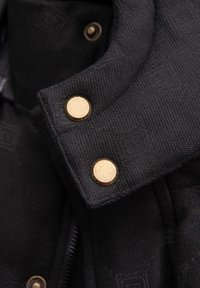 Svart texturerad jacka med en strukturerad krage, guldknappar och en synlig dragkedja. Subtilmönstrad detalj på tyget.