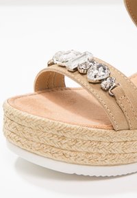 Sandale à plateforme beige avec semelle en corde tressée et bride ornée de grosses pierres décoratives transparentes.