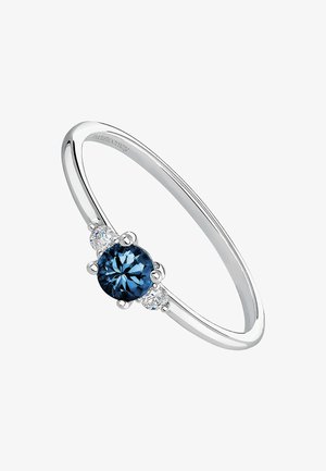 Anillo de plata con una piedra redonda azul central, flanqueada por dos piedras claras más pequeñas, y con una banda lisa y pulida.