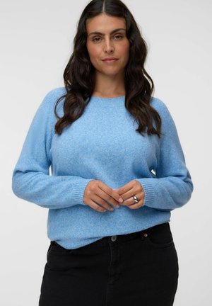 Femme aux longs cheveux foncés portant un pull bleu clair et un pantalon noir, tenant doucement ses mains devant elle sur un fond uni.