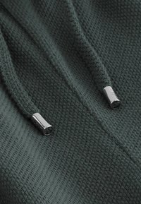Dark green knitted fabric with a textured surface, featuring silver metal aglets on drawstrings for closure.