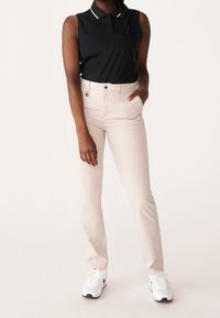 Žena oblečená v světle béžových slim-fit kalhotách, černém bezrukávovém polo tričku a bílých teniskách, stojící s jednou rukou v kapse na bílém pozadí.