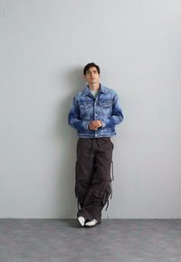 Light blue denim jacket, gray shirt, dark cargo pants with pockets, white sneakers. Standing pose against a plain gray background.