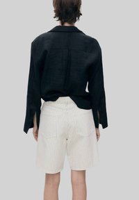Zwarte linnen overhemd met knoopdetails, gecombineerd met witte gestreepte shorts met twee achterzakken. Neutrale achtergrond.