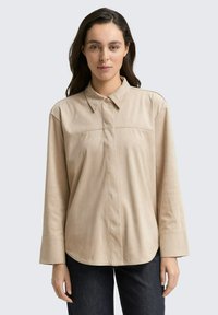 Camisa beige de manga larga con cuello, que presenta un diseño frontal limpio, una textura suave y un corte relajado. Combinada con jeans oscuros.