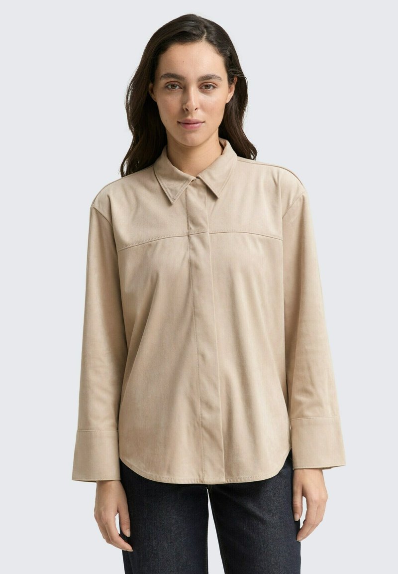 Beige Langarmhemd mit Kragen, das ein schlichtes Frontdesign, eine weiche Textur und einen lockeren Schnitt aufweist. Kombiniert mit dunklen Jeans.