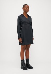 Vestido envelopado em cetim preto com mangas longas e gola, apresentando uma saia ligeiramente evasê. Usado com botas pretas robustas de fivela para contraste.