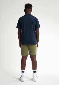 Chemise bleu marine à manches courtes, shorts vert olive, chaussettes blanches avec des rayures noires et baskets blanches, vues de dos.