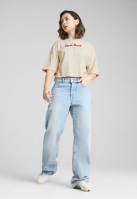 Beige cropped t-shirt med rød "Puma Resort" tekst, parret med lyseblå løse jeans og lyse sko. Ensfarvet baggrund.