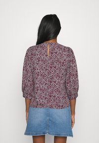 Femme aux longs cheveux noirs portant un haut bordeaux à fleurs avec des manches trois-quarts et une jupe en jean bleu clair, vue de dos.