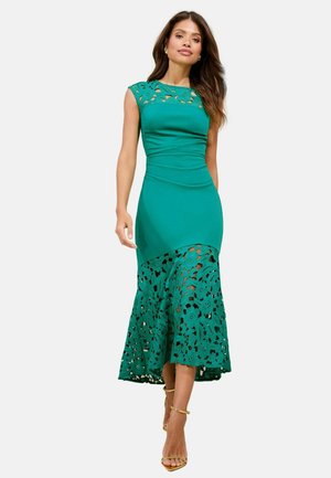 Žena v zelených priliehavých šatách bez rukávov s čipkovanými detailmi na hornej časti hrudníka a dolnej časti sukne, v kombinácii so zlatými sandálmi na vysokom podpätku.