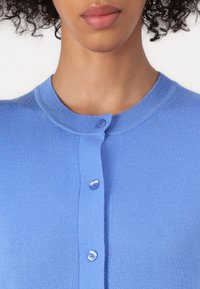 Cardigã azul claro com gola redonda, feito de um tecido suave, textura canelada e cinco botões combinando na frente.