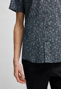 Camicia a maniche corte in tessuto scuro con un motivo floreale blu. Presenta una chiusura a bottoni sul davanti e una vestibilità comoda, abbinata a pantaloni neri.