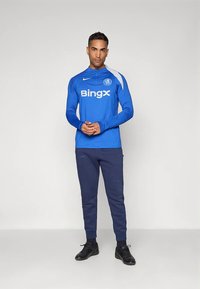 Muž v modrom tréningovom tričku Chelsea FC a tmavých teplákoch stojí so zloženými rukami na pozadí bielej steny.