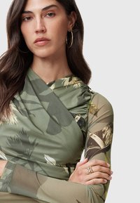 Top a maniche lunghe verde con motivo floreale, realizzato in tessuto drappeggiato e con accenti trasparenti; modello che indossa orecchini a cerchio dorati e anelli sovrapposti.