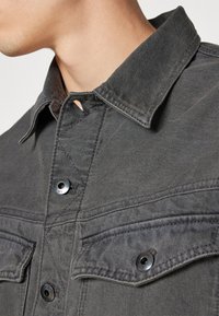 Γκρι τζιν πουκάμισο με αιχμηρό γιακά, κουμπωτή μπροστινή πλευρά και δύο τσέπες στο στήθος με μεταλλικές λεπτομέρειες. Διακριτή υφή ορατή στο ύφασμα.