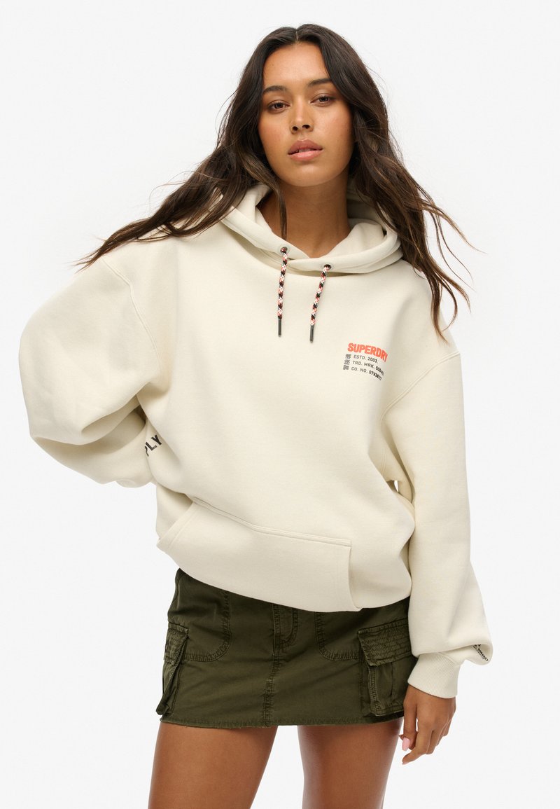 Sweatshirt à capuche crème avec une poche avant, doté de cordons noirs et rouges et d'un impression de logo ; associé à une jupe cargo verte.
