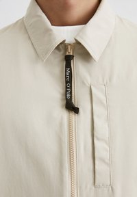 Beige lightweight jacket with a zip front, stand collar, and branded drawstring. Features a side pocket with a textured fabric.