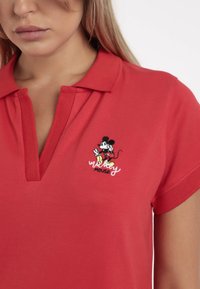 Camisa polo roja de algodón con un acabado texturizado, que presenta un gráfico bordado de Mickey Mouse y un sutil detalle en el cuello.