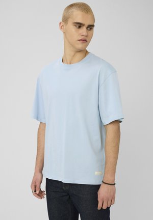 Jeune homme aux cheveux blonds courts, portant un t-shirt oversize bleu clair et un jean foncé, regardant vers sa gauche sur un fond gris uni.
