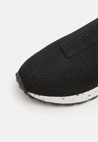 Bout de sneaker en mesh noir avec semelle blanche tachetée et étiquette en tissu noir marqué sur le dessus.
