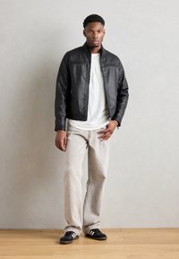 Jachetă din piele artificială neagră cu detalii quilted, asortată cu un tricou alb, blugi bej deschis și adidași negri cu dungi albe.