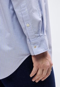 Κοντινό πλάνο ενός χεριού δίπλα στο μανίκι ενός μπλε και άσπρου ριγέ πουκαμίσου με λευκά κουμπιά, φορεμένο πάνω από σκούρο παντελόνι.