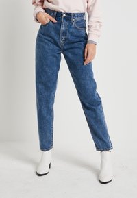 Högmidjade blå denimjeans med rak ben, fem fickor och metalldetaljer. Matchas med vita klackiga ankelstövlar.