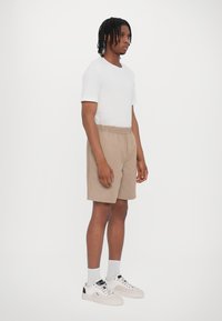 Bruna shorts med resårmidja, åtföljda av en figurnära vit t-shirt. Modellen bär grå strumpor och vita sneakers med svarta detaljer.