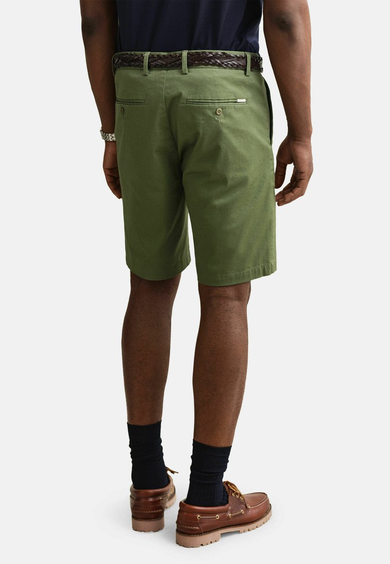 Hombre con pantalones cortos hasta la rodilla color verde oliva, cinturón trenzado marrón, calcetines negros y zapatos de cuero marrones, de pie contra un fondo blanco.