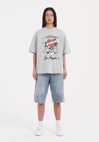 Camiseta gris de gran tamaño con estampado gráfico que presenta una calavera y un corazón, combinada con pantalones cortos de mezclilla azul claro y zapatillas blancas.