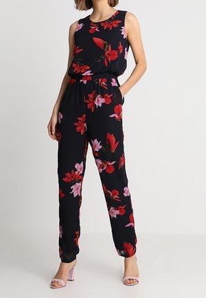 Combinaison noire sans manches avec imprimé floral rouge et rose, taille élastique, poches latérales, associée à des talons ouverts rose clair.