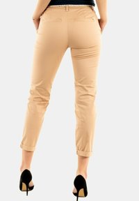 Pantalons ajustés beige clair avec un ourlet roulé, deux poches arrière et une texture lisse, associés à des chaussures à talons hauts noires.