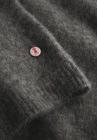 Donkergrijze wollen trui met een opvallende zilveren metalen badge met een rode dieren-silhouet en een klein rood lint. Zacht, textuur voelend stof.