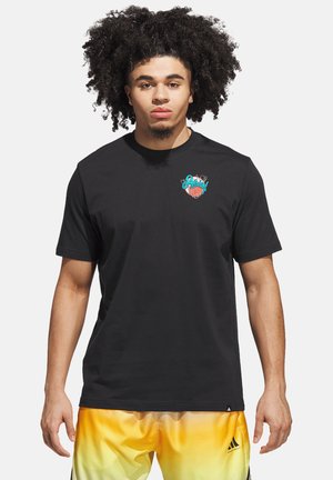 Joven con cabello rizado que lleva una camiseta negra con un pequeño logo colorido de baloncesto y pantalones cortos deportivos en degradado amarillo.