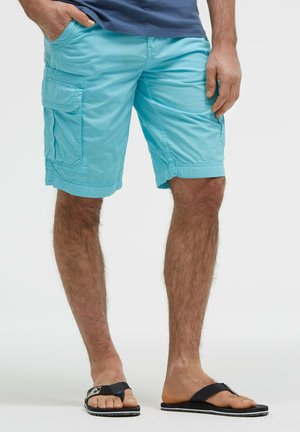 Male wearing light blue cargo shorts, blue shirt, and black flip-flops, standing with one hand in pocket against white background.
