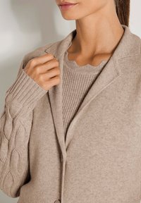 Frau trägt einen beigen Strickpullover mit gewelltem Ausschnitt unter einem beigen Kabelstrick-Cardigan und hält den Cardigan-Kragen nahe der Schulter.