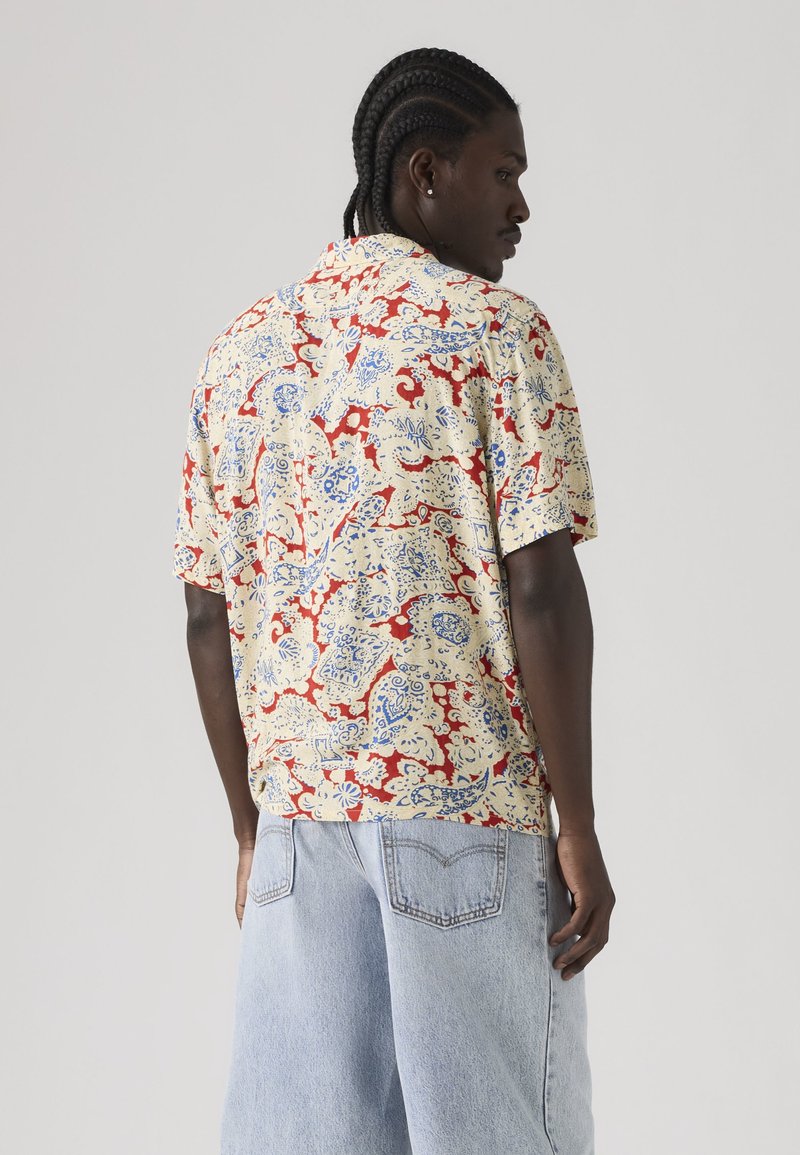 Mand med flettet hår iført en kortærmet beige skjorte med røde og blå paisley-mønstre og lyseblå denimshorts, der vender sidelæns.