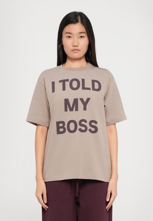 Beige t-skjorte med rund hals og korte ermer, med fet svart tekst: "JEG FORTALT TIL SJEFEN MIN." Stoffet ser mykt ut, med en avslappet passform.