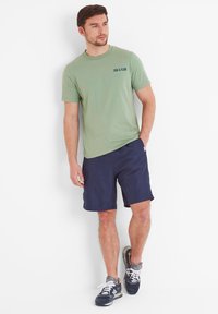 Green cotton t-shirt with "EBB & FLOW" text, paired with navy shorts and blue sneakers featuring white laces and grey accents.